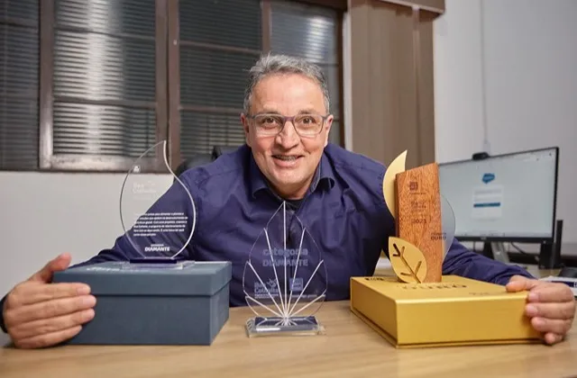 Homem sorrindo e segurando troféus sobre a mesa